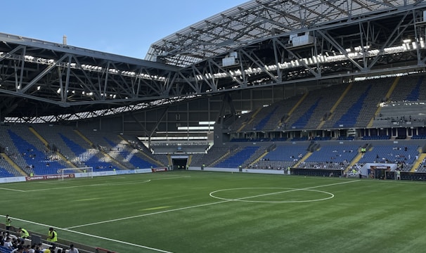 A modern sports stadium with a large, green soccer field and surrounding seating areas. Blue and yellow seats are visible, with some spectators scattered across the stands. The stadium is partially covered with a steel lattice roof, showing a clear blue sky. Several security personnel in high-visibility vests are present.
